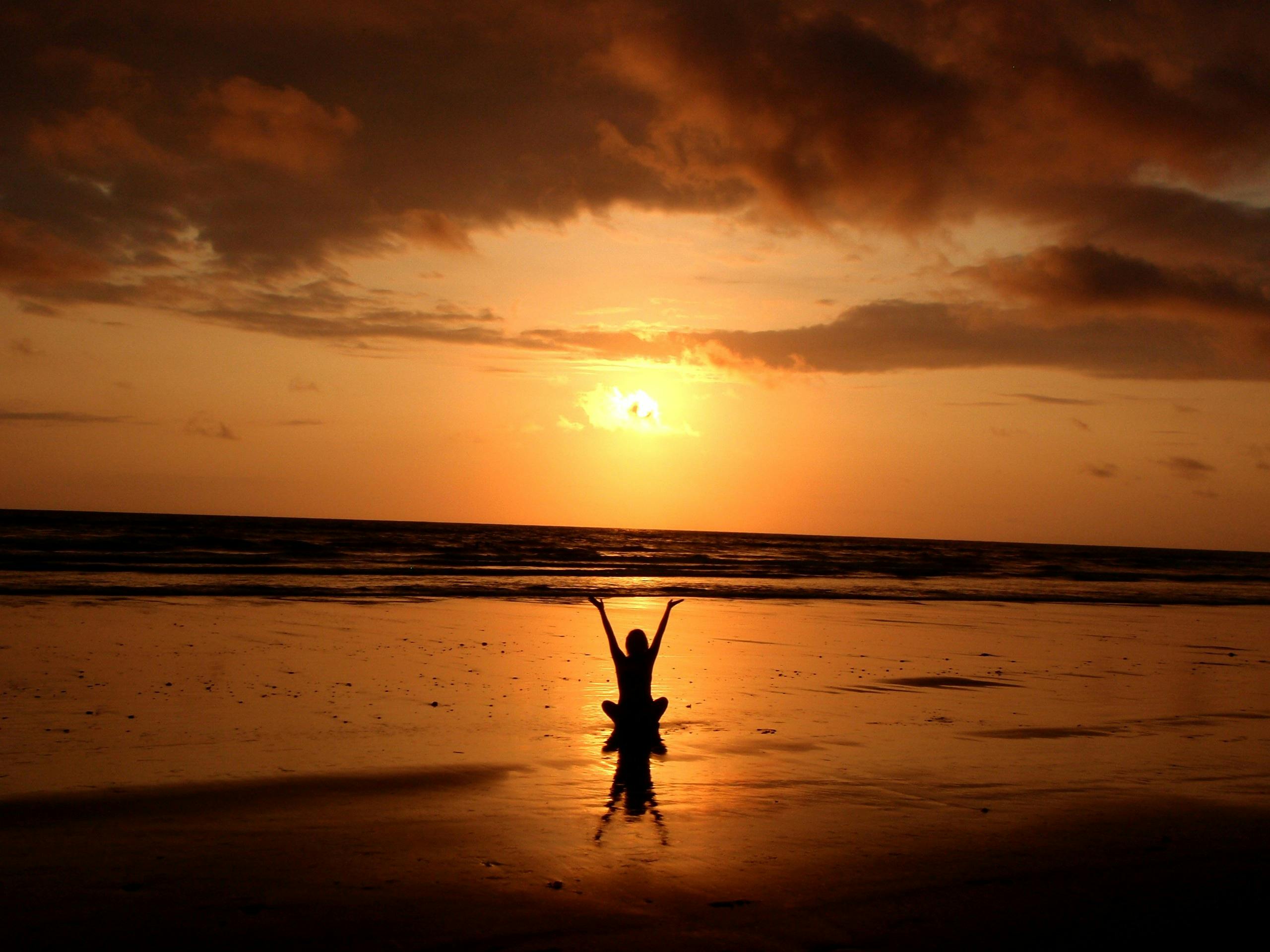 Silhouette of a person raising their hand during sunset on a serene beach.

